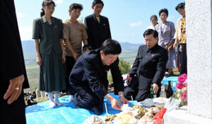 people in formal clothing place and arrange food and drinks on a blue tarp at a hillside memorial with others standing nearby against a rural landscape